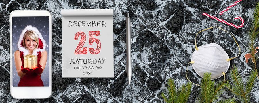 tear-off calendar with the date of christmas day 2021 and a smartphone with picture of a smiling miss santa holding a golden present on stone pavement background