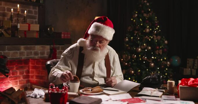 Santa Claus eating ginger cookies, dipping into milk close-up. Mr Claus wearing circle glasses relaxing after work, sitting at workplace in cozy house, free time. Post office of Santa, leisure.