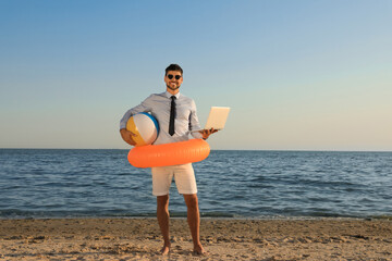 Happy man with inflatable ring, ball and laptop near sea on beach. Business trip