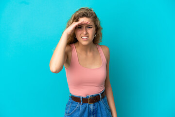 Young blonde woman isolated on blue background looking far away with hand to look something