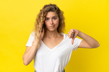 Young blonde woman using mobile phone isolated on yellow background showing thumb down with negative expression