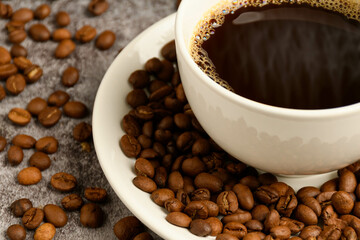 Close-up, Crop, Selective focus of coffee beans are in coffee cup saucer. Hot coffee in a white cup is placed on a cement floor table in an office, smoke and aroma waft from a cup. Blurred background