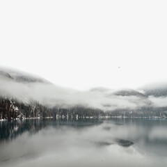 Mountain lake in winter. Minimalist landscape in black and white colors. Amazing panoramic winter natural landscape snow-capped mountain peaks, pine trees reflected in the water of the lake clouds fog