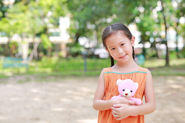 Portrait of happy little Asian child in green garden with hugging teddy bear and looking at camera. Close up smiling kid girl in summer park.