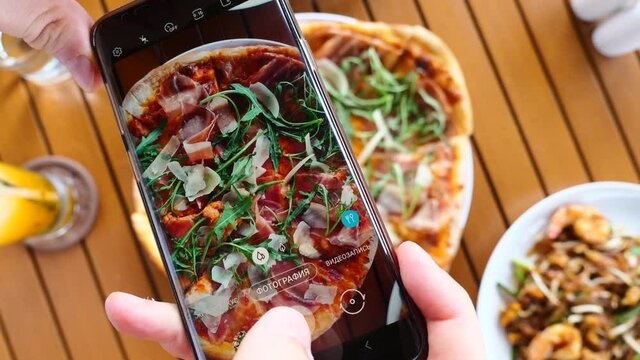 Man's hands hold a cell phone and take photos of Italian food for social media. Blogger man taking pictures of fresh pizza on his smartphone. Technology, food porn concept. Photography Word on Russian