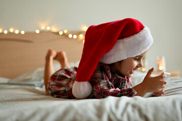 Smiling little girl in a Red Santa hat making video calls to family and friends. Online Christmas greetings. Cute Child communicates and has fun, using Smartphone. Merry Christmas and Happy New Year.