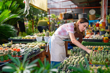 Young Asian woman gardener caring houseplant and flower in greenhouse garden. Female plant shop owner using digital tablet checking plants in store. Small business entrepreneur and plant shop concept