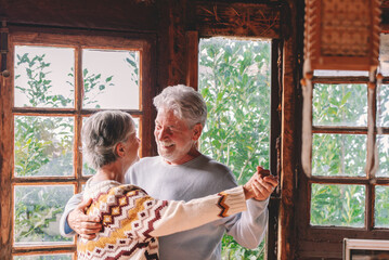 Happy senior couple smile and dance at home enjoying love and relationship together. Active old man and woman have fun in indoor leisure activity. Nature woods view from windows - Powered by Adobe