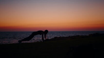 Strong young woman in silhouette standing in plank position during workout on ocean beach. Female athlete in sport outfit training abs with colorful summer sunset on background.