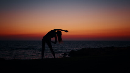 Young woman with slender body practising in fitness exercises during evening time near ocean. Silhouette of active healthy female. Regular training for body care.