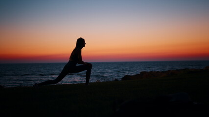 Silhouette of fitness young woman stretching legs after active workout on beach. Beautiful summer sunset over ocean. Concept of healthy and active lifestyles.