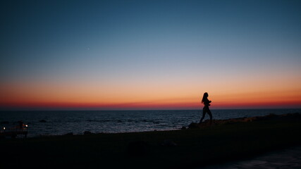 Obraz premium Silhouette of sporty young woman in activewear running along beach during evening time. Physical exercises during summer sunset over ocean. Motivation and endurance concept.