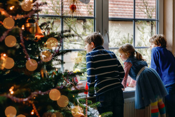 Three cute children sitting by window on Christmas eve. Two school kid boys and toddler girl,...