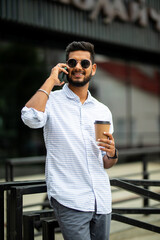 Business talk on a go. Confident young indian man in glasses holding coffee cup and talking on mobile phone while walking outdoors