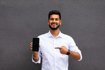 Portrait of cheerful, positive, attractive man having smart phone with black screen in hand, pointing with forefinger to product, isolated on grey background