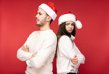 Obraz premium Photo of attractive couple in Christmas hats, stands back to back and looks resentful and desperate, isolated over red background