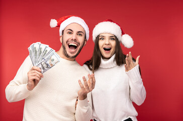 Photo of sweet couple in Christmas hats, holds money in dollars, sales. Male and female are in love, look blessed and smile, isolated over red background