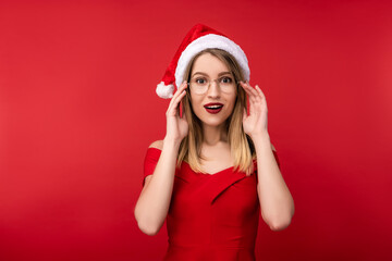 Photo of attractive woman in Christmas hat and red dress, holds glasses with surprised. Isolated over red background