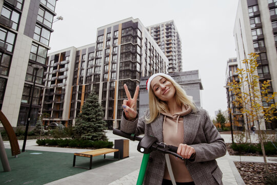 Posing for photo Young cheerful blonde woman showing peace sign and looking at camera with smile. Grey modern apartment blocks on background. She driving electical scooter.