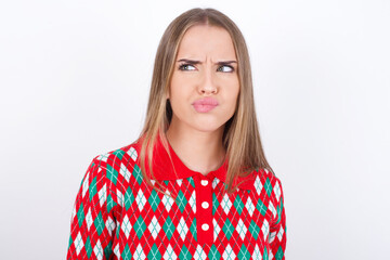 Young caucasian girl wearing christmas sweaters on white background crosses eyes, puts lips, makes grimace with awkward expression has fun alone, plays fool.