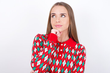 Portrait of thoughtful Young caucasian girl wearing christmas sweaters on white wall keeps hand under chin, looks away trying to remember something or listens something with interest. Youth concept.