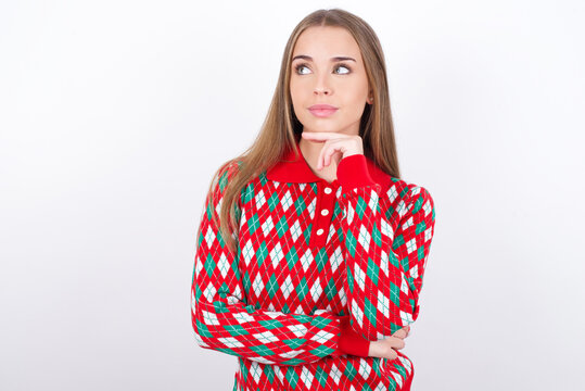 Dreamy Young caucasian girl wearing christmas sweaters on white background with pleasant expression, looks sideways, keeps hand under chin, thinks about something pleasant.