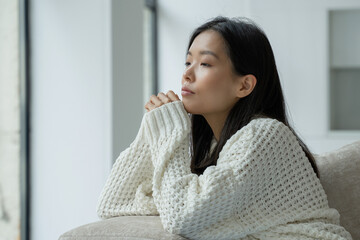 A young unhappy Asian woman sitting on the couch with a sad face and looking out the window.