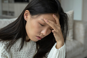 Hand on the temples of a young sad Asian woman sitting on the sofa. She feels bad because of the headache.