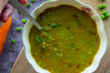Eating Homemade pea soup with a spoon that is rich in flavors, vegetables, and healthy ingredients. Hot green split pea soup for winter in a decorative bowl served on a wooden board. Fresh and tasty.