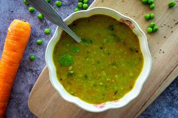 Homemade green pea soup with a spoon. Hot split pea soup rich in flavors, vegetables, herbs and healthy ingredients. Tasty soup for winter in a decorative bowl served on a wooden board. Top view