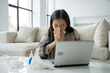 An Asian woman blows her nose in a napkin, sits at a home office desk, studies on a laptop, a sick woman has the flu, covered herself with a blanket, caught a cold