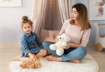 A middle-aged mom is playing with her little daughter. in the hands of a woman and a girl a white and brown bear. Beautiful delicate photo in pink tones