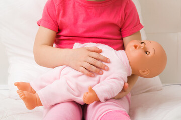 Toddler girl sitting on bed, holding and putting to sleep baby doll on lap. Front view. Closeup....