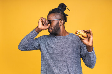 Bad smell. Young african american black man eating hamburger isolated over yellow background. Diet...