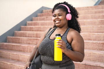 Happy plus size African woman doing workout routine while listening music with wireless headphones...