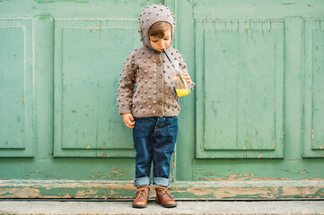 Outdoor portrait of cute toddler girl wearing brown knitted jacket, drinking orange juice from the...