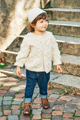 Outdoor portrait of adorable toddler girl wearing white knitted pullover and hat, dark blue denim jeans and brown retro shoes, fashion for little children