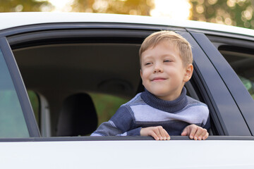 A seven year old cute boy leans out the window of a white car on a warm sunny autumn day against the backdrop of yellow foliage. Selective focus. Portrait