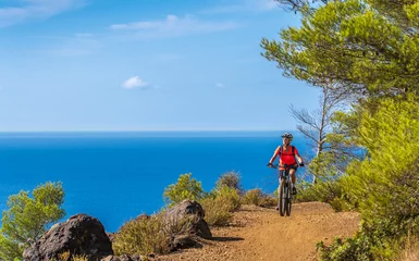 Tuinposter nice woman riding her electric mountain bike at the coastline of mediterranean sea on the Island of Elba in the tuscan Archipelago, in front of Porto Ferraio,Tuscany, Italy © Uwe