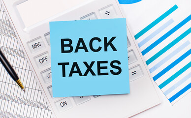 On the desktop is a white calculator with a blue sticker with the text BACK TAXES, a pen and blue reports. Business concept