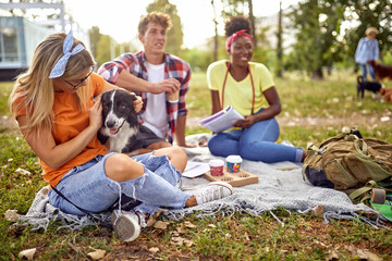 A group of students are sitting on the grass in the park and enjoying coffee. Friendship, rest, pets, picnic