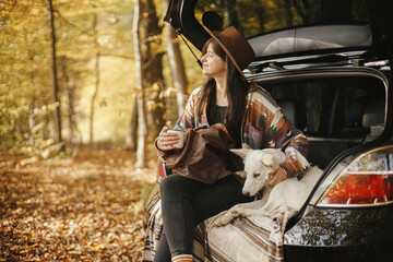 Stylish woman traveller with backpack sitting with cute dog in car trunk in sunny autumn woods. Young hipster female traveling with swiss shepherd white dog. Travel and road trip with pet