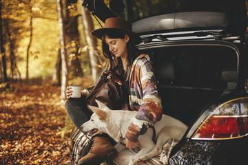 Stylish hipster woman with cup and backpack sitting with cute dog in car trunk in sunny autumn woods. Travel and road trip with pet. Space for text. Young female traveler hugging sweet white dog