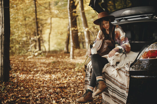 Stylish hipster woman with cup and backpack sitting with cute dog in car trunk in sunny autumn woods. Travel and road trip with pet. Space for text. Young female traveler hugging sweet white dog