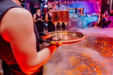 Handsome waiter holding in naked hands tray with glasses full of champagne. Night party celebration in club with colour disco lights. Professional restaurant serving in banquet, luxury event.