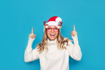 Fototapeta na wymiar A young amazed blonde woman in glasses with deer antlers, a white warm sweater and Christmas Santa hat points up to empty copy space for greeting text with fingers isolated on color blue background