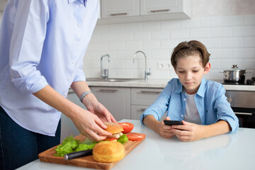 Mom is in the kitchen preparing a homemade burger for her teenage son..An 11-year-old boy is sitting at the kitchen table and looking at his mobile phone.