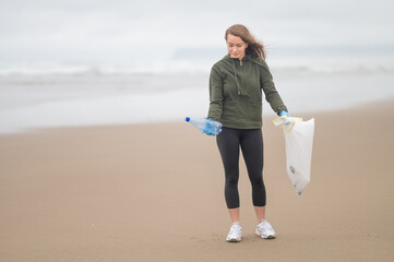 Girl volunteer in sportswear collects used plastic bottles on the ocean shore. Sandy shore,...
