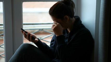 Woman rubbing eyes and face with hand while holding cellphone by window.
