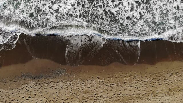 Drone view of beautiful endless images in ultra slow motion as the foamy waves break on the sandy shore. Aerial shot of a golden beach meeting deep ocean waters.